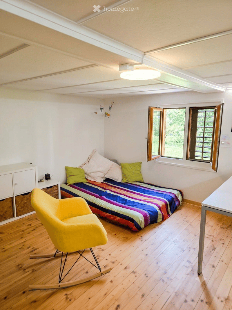 Cozy bedroom with wooden flooring, a single bed with colorful striped bedding, a yellow rocking chair, a small cabinet, and a table near the windows.