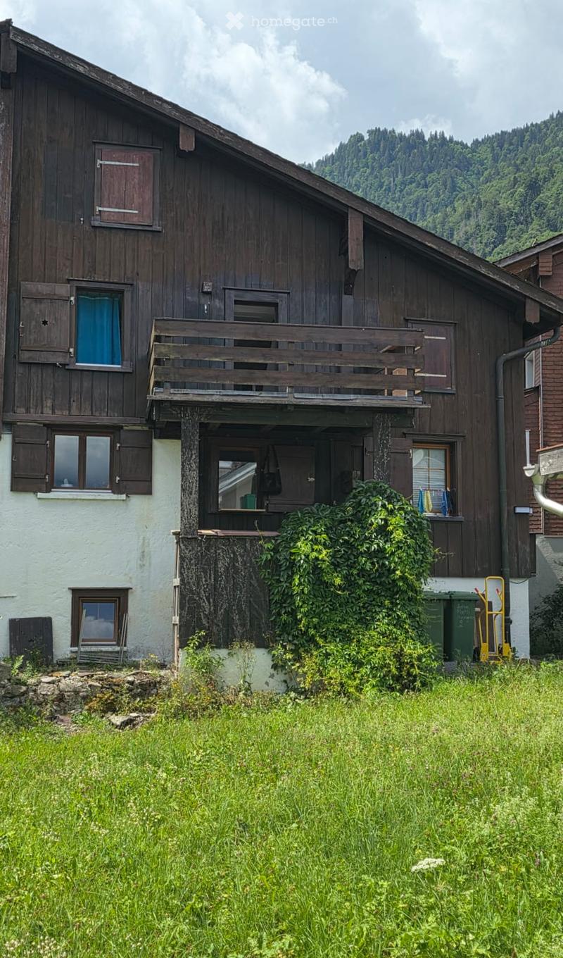 A two-story wooden house with a balcony, multiple windows, and a surrounding garden area.