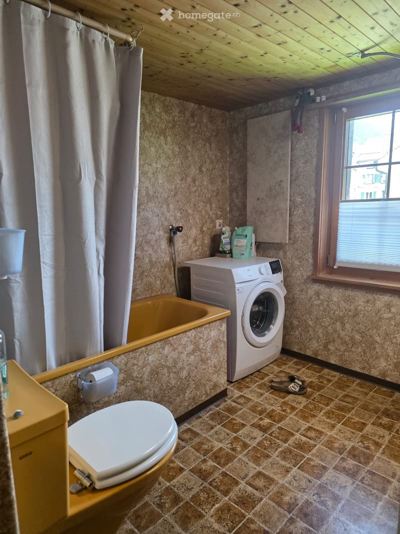 Bathroom with a toilet, bathtub, washing machine, and shower curtain. The room features tiled walls and a window with blinds.