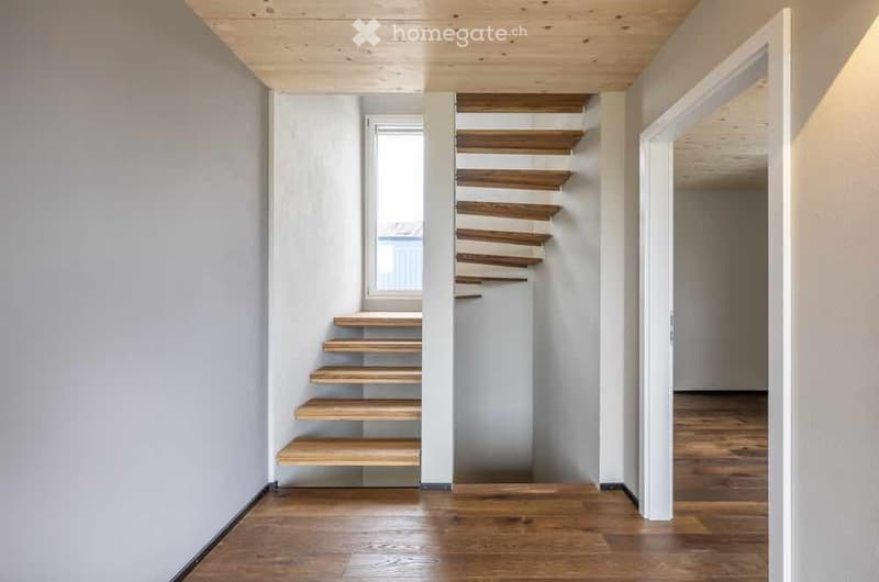 Narrow hallway with wooden stairs leading upwards, light grey walls, and wooden flooring.