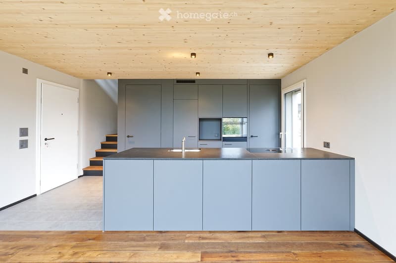 Modern kitchen with grey cabinets, a central island, and a wooden floor. Features a sleek sink and faucet, with built-in storage and a raised ceiling with recessed lighting.