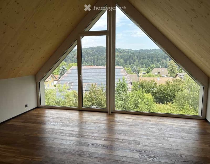 Bright, empty attic room with wooden flooring and large, angled windows offering a scenic view of the surrounding landscape.