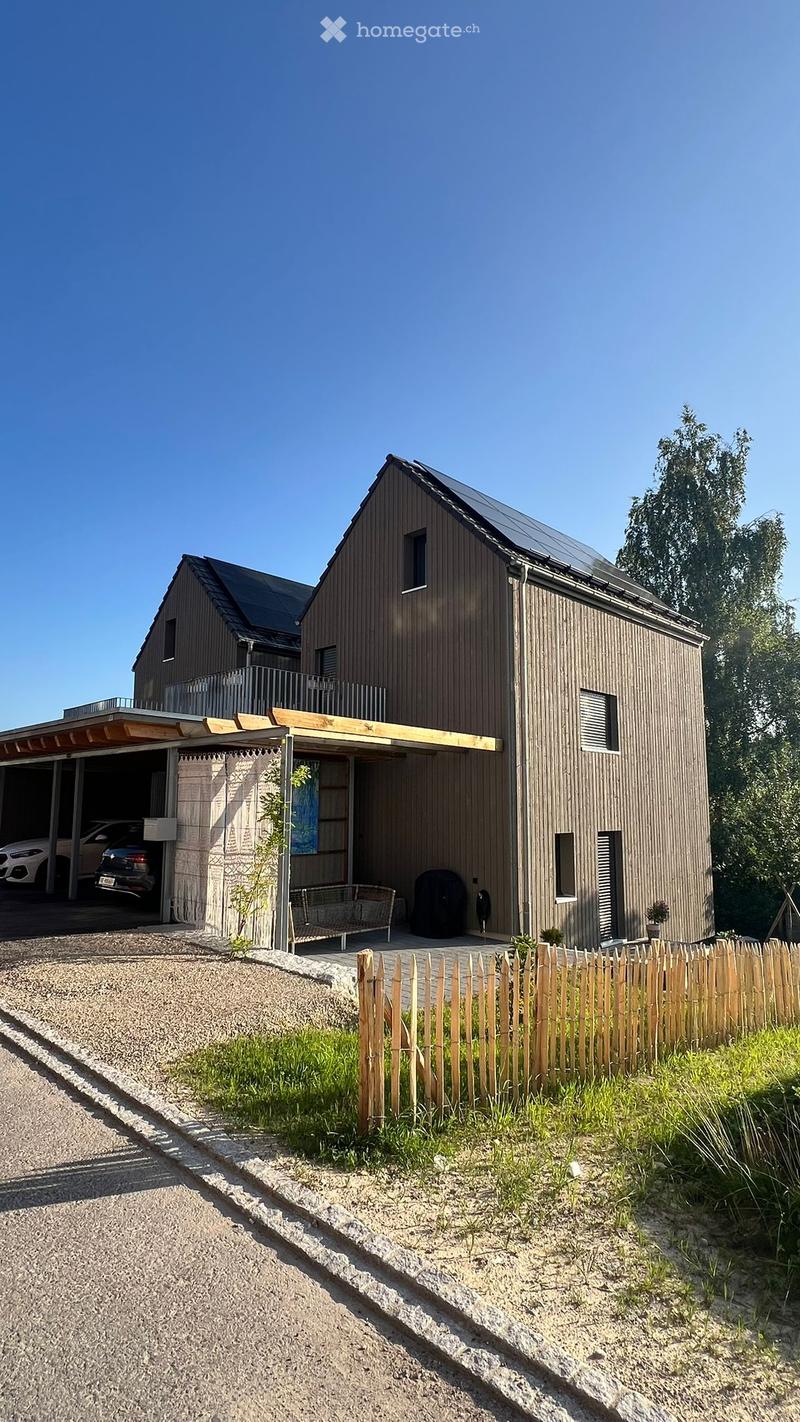 Modern two-story house with wooden siding, solar panels on the roof, a covered garage, and a small outdoor seating area.