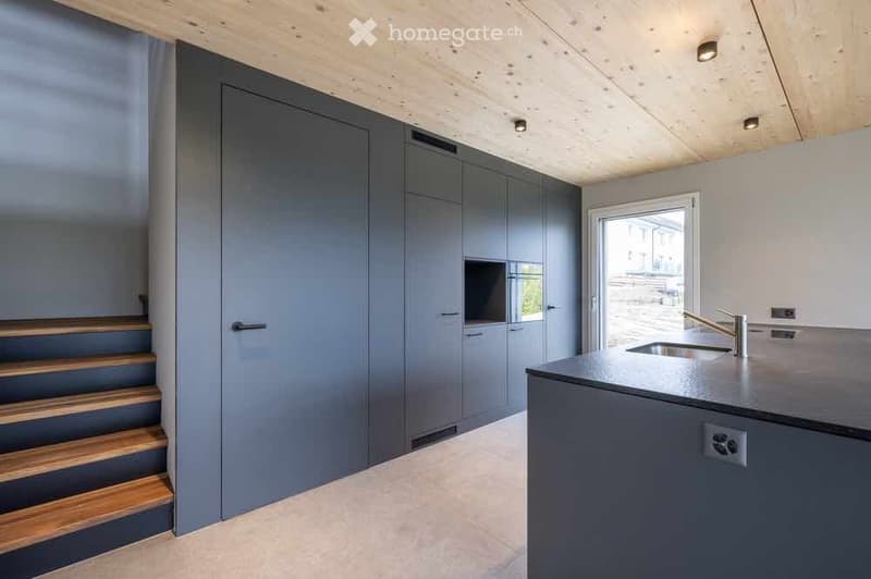 Modern kitchen with grey cabinetry, a black countertop, and a stainless steel sink. Wooden staircase leads upwards.
