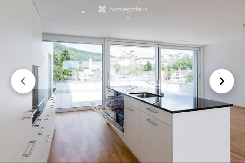 Modern kitchen with white cabinets, black countertop, stainless steel sink, and dishwasher. Large windows offer a scenic view of the city and mountains.
