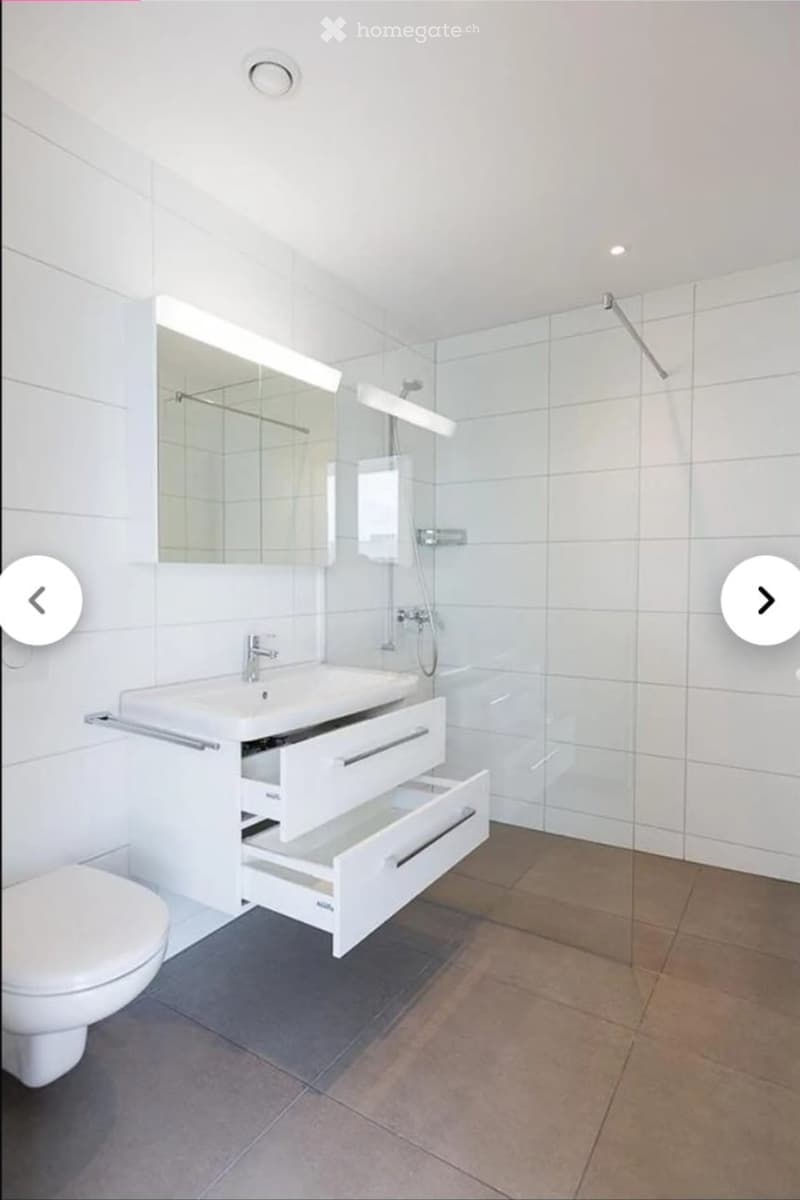 Modern bathroom with white tiles, a sink with a cabinet, a toilet, a mirror, and a glass-enclosed shower area.