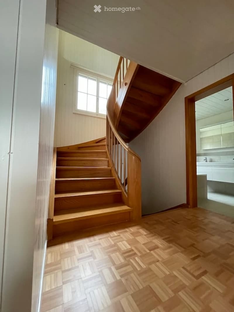 Spacious hallway with wooden flooring and a wooden staircase leading to an upper level. A window provides natural light, and a partially visible bathroom is seen to the right.