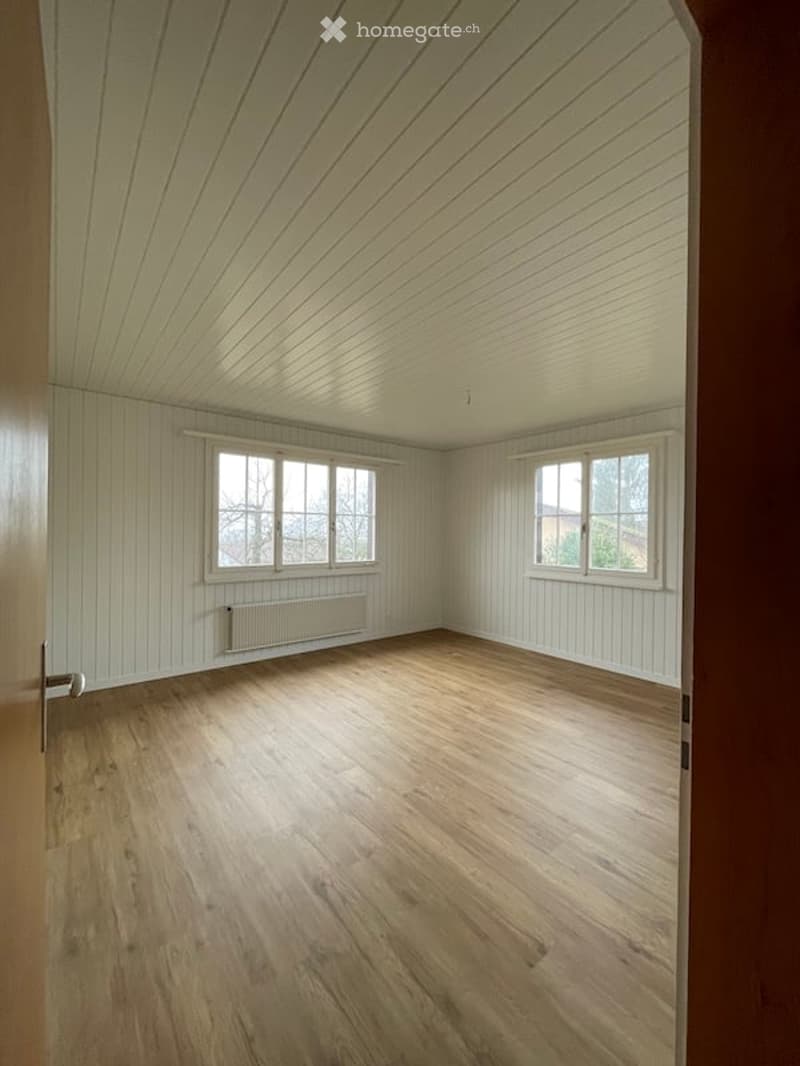 Empty room with wooden flooring, white paneled walls, and two windows with white frames. The ceiling has a sloped design with wooden paneling. A radiator is visible below one of the windows.