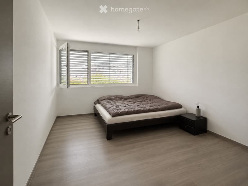 Minimalist bedroom with a single bed, nightstand, and lamp. The room features light wooden flooring and white walls, with a window offering a view of greenery outside.