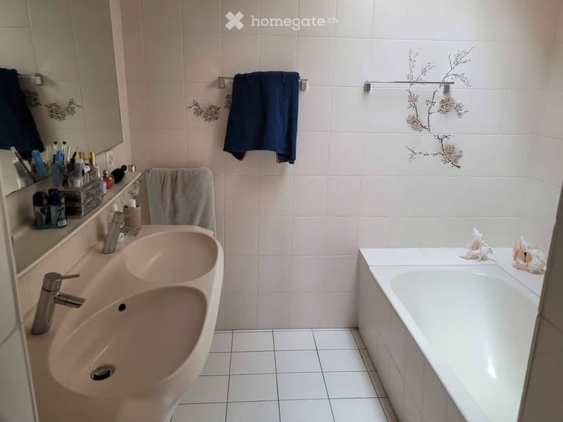 A modern bathroom with white tiled walls and floor, featuring a double sink, a bathtub, a mirror, and decorative towels.