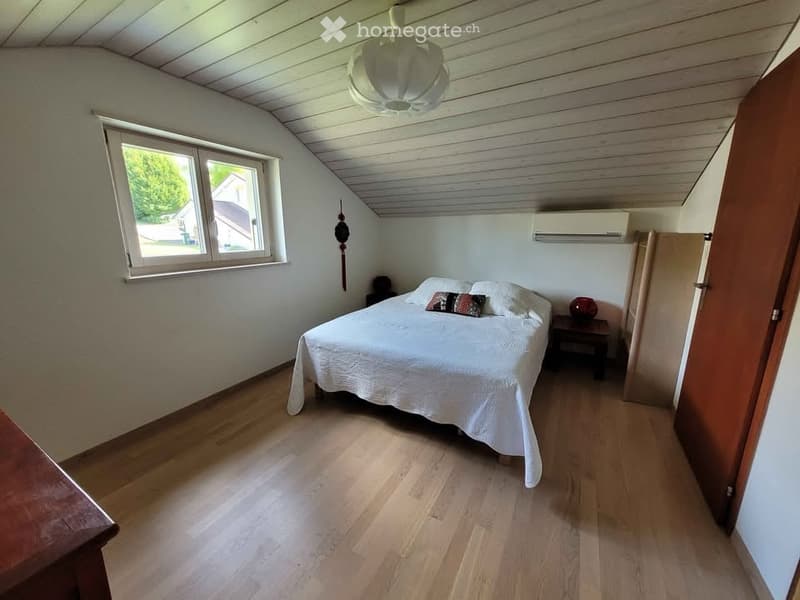 Cozy attic bedroom with wooden floors, slanted ceiling, large window, bed with white bedding, nightstand, and decorative items.