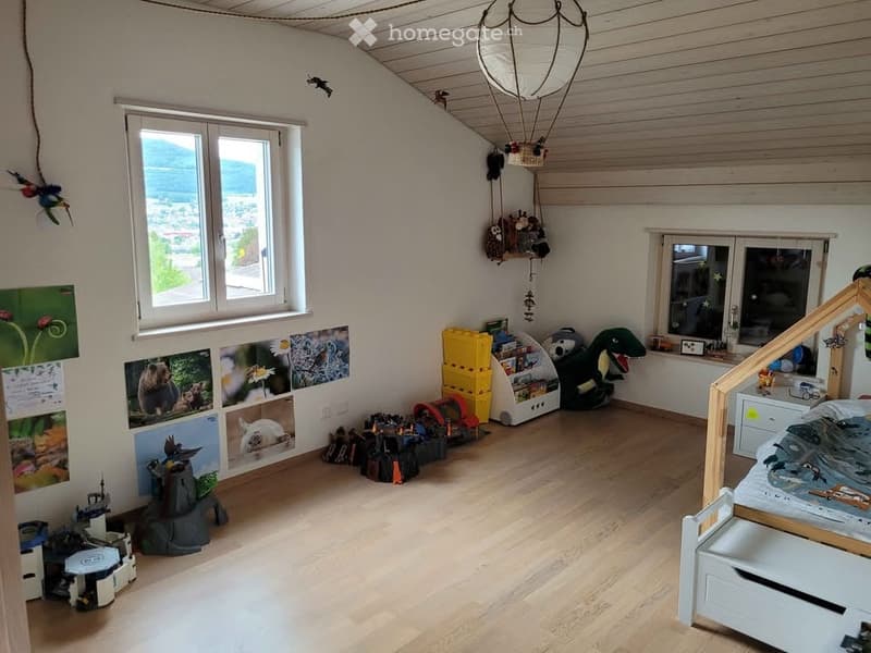 Bright, airy children's room with wooden flooring, large windows offering a scenic view, a bed with a themed bedspread, a colorful toy box, a bookshelf, a plush toy dinosaur, and various toys and decorations including picture frames and a balloon decorati