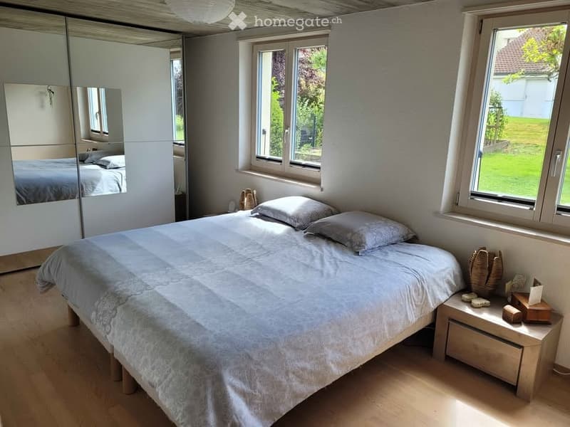 Bright bedroom with a double bed, nightstand, mirror, and wardrobe. Large windows provide natural light and a view of the garden.