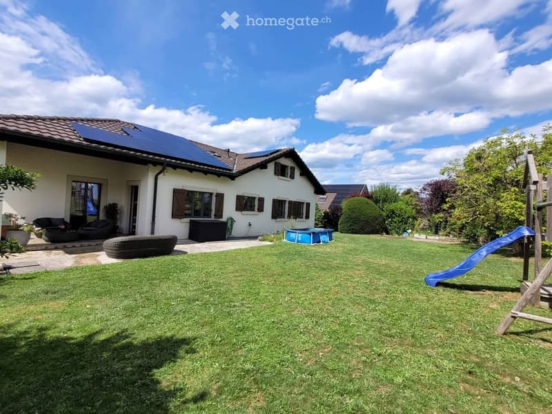 A well-maintained garden with a large lawn, a blue plastic pool, a blue slide, and outdoor seating including a black wicker sofa and a round black ottoman. The house in the background is white with solar panels on the roof and several windows.