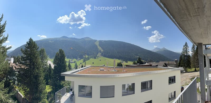 Modern white building with a flat roof and multiple windows, overlooking a scenic mountain landscape with ski slopes and forested areas.