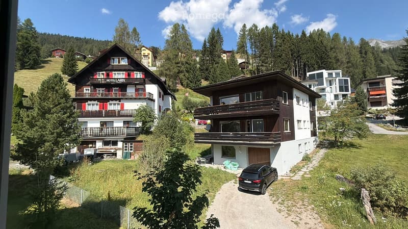 A scenic view of a multi-story wooden house with balconies and red shutters, surrounded by lush greenery and other houses in a mountainous area.