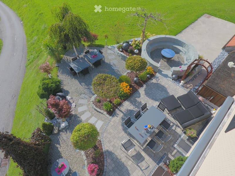 Well-maintained garden with a patio area, outdoor dining table, lounge chairs, and a circular fire pit surrounded by gravel and greenery.