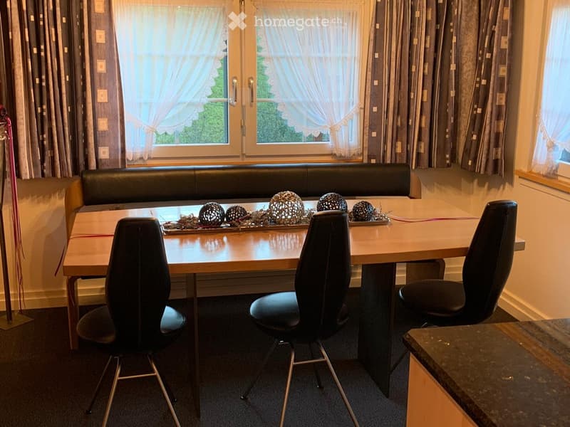 A small, cozy dining area with a wooden table, black chairs, and a built-in leather sofa bench. The room features large windows with curtains and a decorative centerpiece on the table.