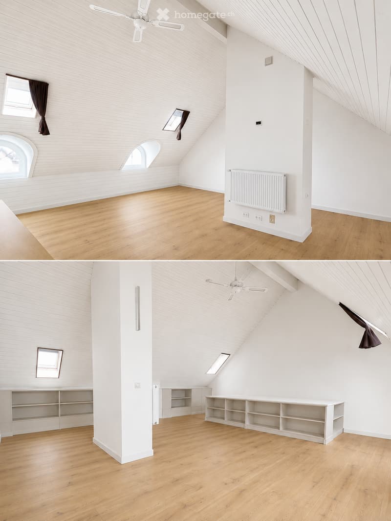 Bright, empty attic room with wooden flooring, white walls, exposed wooden beams, and a ceiling fan. Features large windows with dark curtains, built-in shelving units, and a radiator.