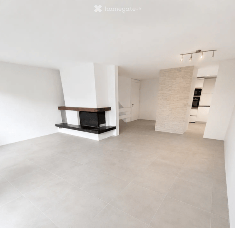 Open-plan living space with a fireplace, white walls, grey tiled floor, and recessed lighting. The room is unfurnished, showing only the fireplace and a doorway leading to another area.