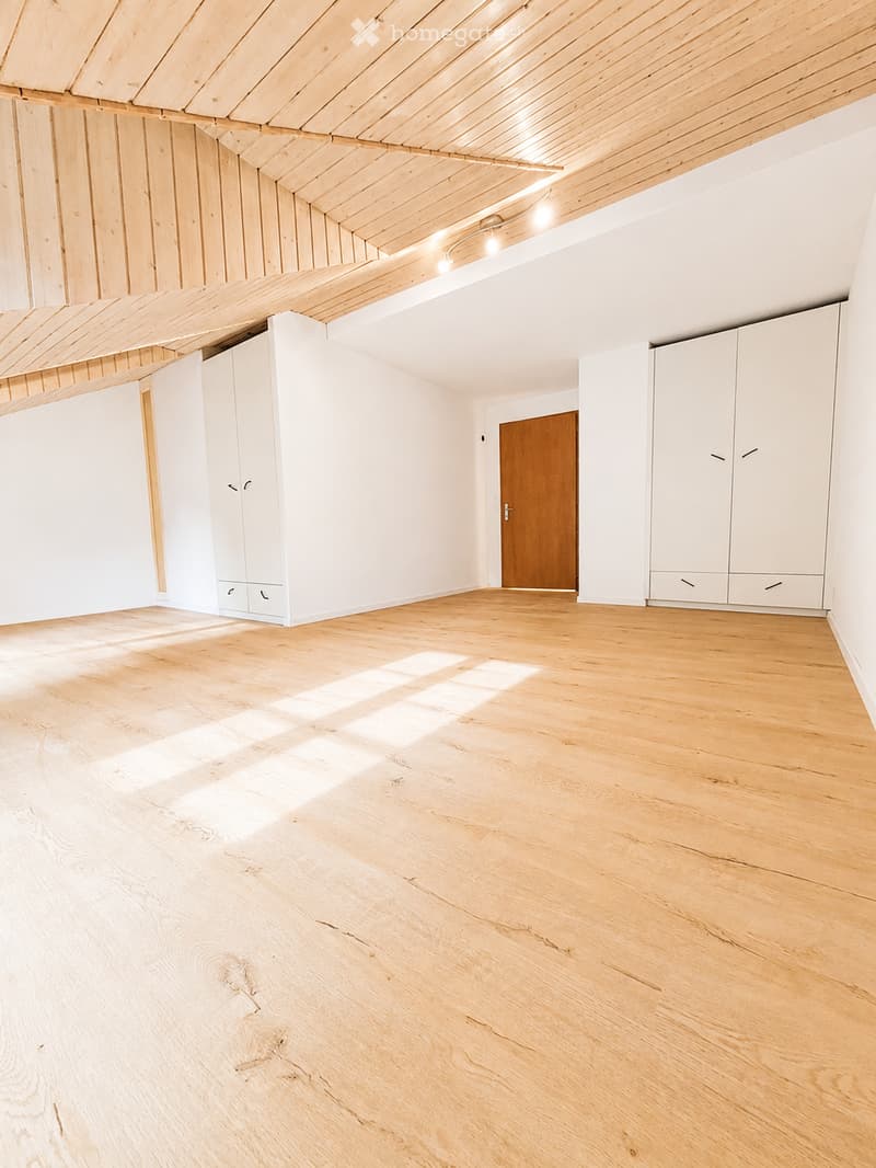Empty room with light wooden flooring, white walls, built-in white cabinets, and a wooden door. The ceiling features exposed wooden beams and integrated lighting.