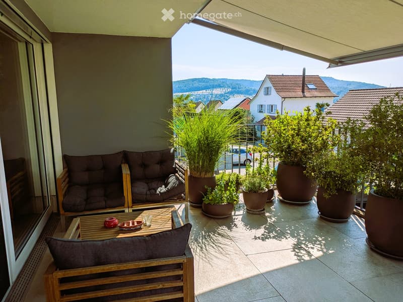 Balcony with a sofa, wooden table, chair, and several potted plants, offering a scenic view of the surrounding area.