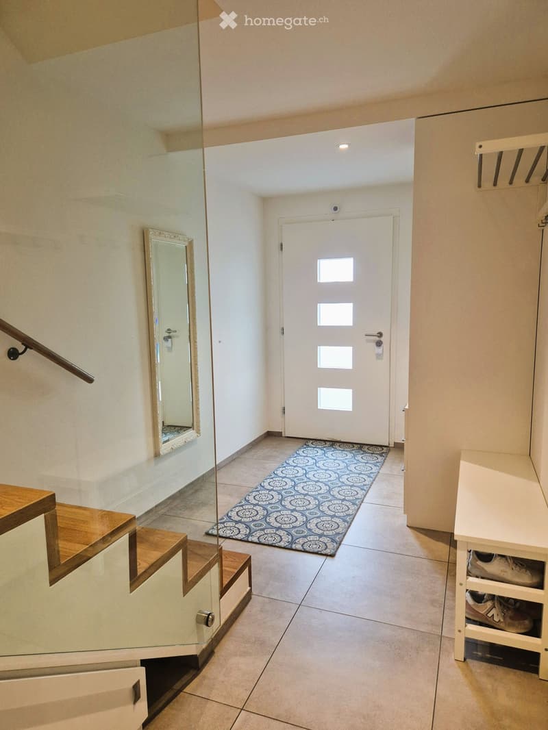 Modern hallway with tiled floor, glass staircase with wooden steps, white door with four windows, patterned rug, and shoe rack with shoes.