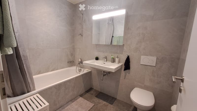 Modern bathroom with a bathtub, sink, toilet, and a large mirror with built-in lighting. The walls are tiled in a light gray color, and there are towels and small decorative items on the sink counter.