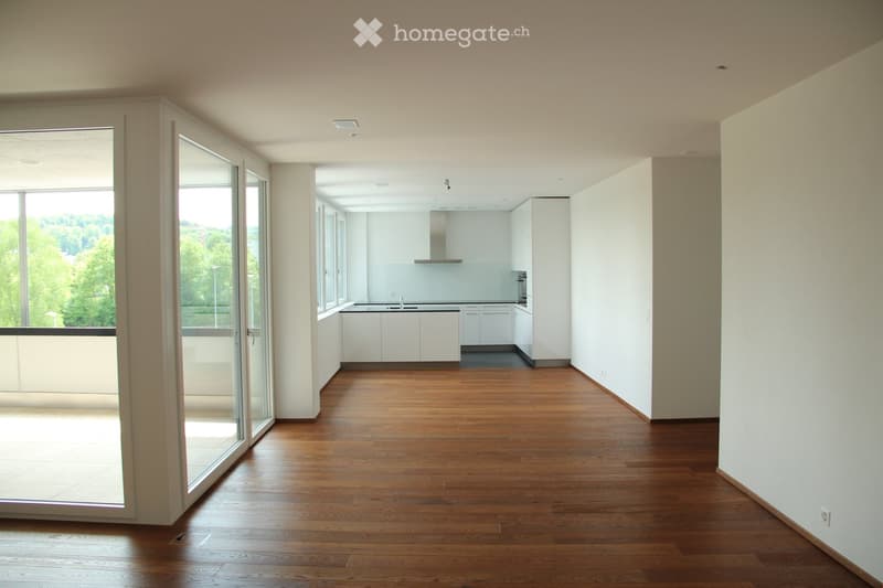 Empty room with wooden flooring, large glass sliding doors leading to a balcony, and a view of greenery outside. The adjacent kitchen area is also visible but unfurnished.
