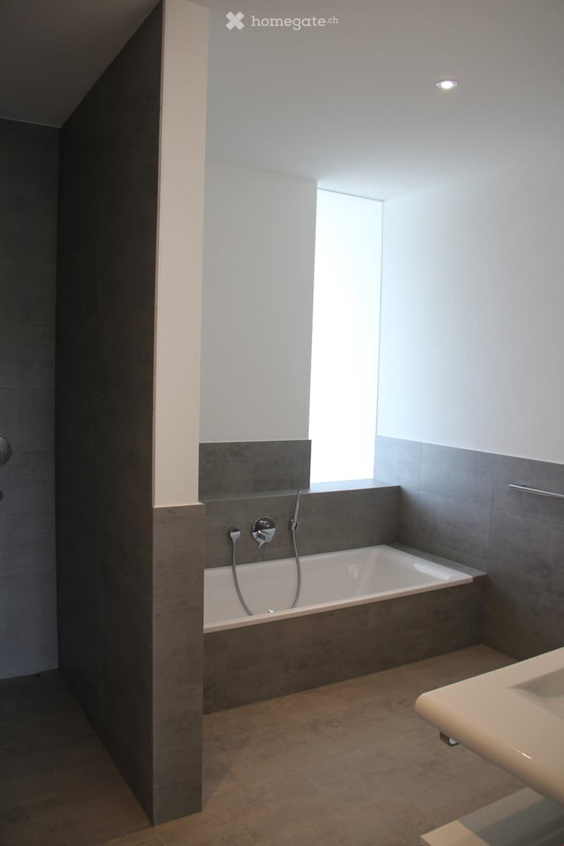 Modern bathroom with a bathtub, faucet, and sink. The walls are tiled in a combination of light and dark gray, and there is a window providing natural light.