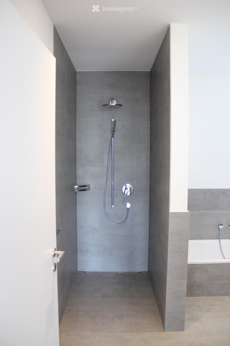 Modern, minimalist bathroom with grey tiles, a shower area, and a bathtub visible in the background.