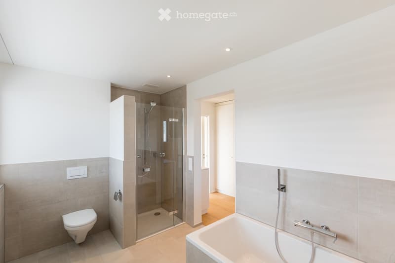 Modern bathroom with a toilet, bathtub, and glass-enclosed shower. Light-colored tiles and wooden flooring.