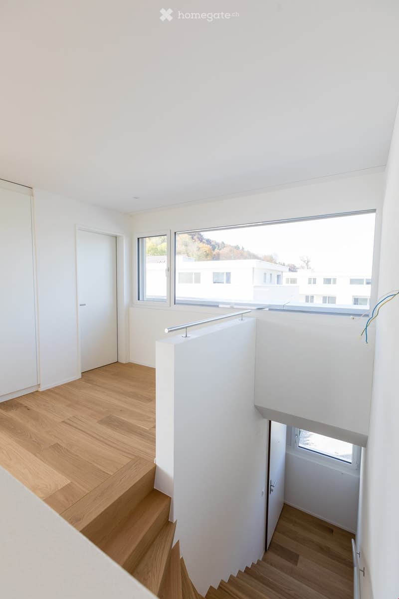 Modern, minimalist hallway with light wooden flooring, white walls, and large windows offering a view of the exterior. Features a staircase with a sleek metal railing and a small landing area.