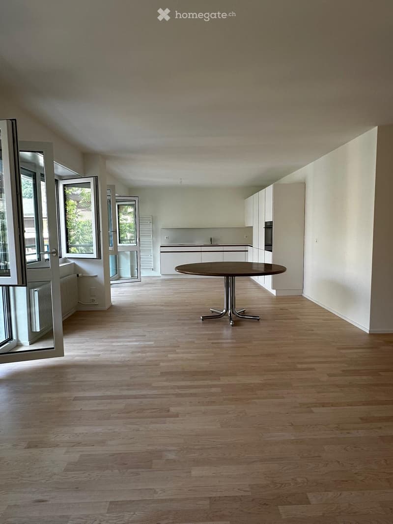 Open-plan space with light wooden flooring, white walls, and a single round table near the kitchen area. Large windows and sliding doors provide natural light and access to the outdoors.