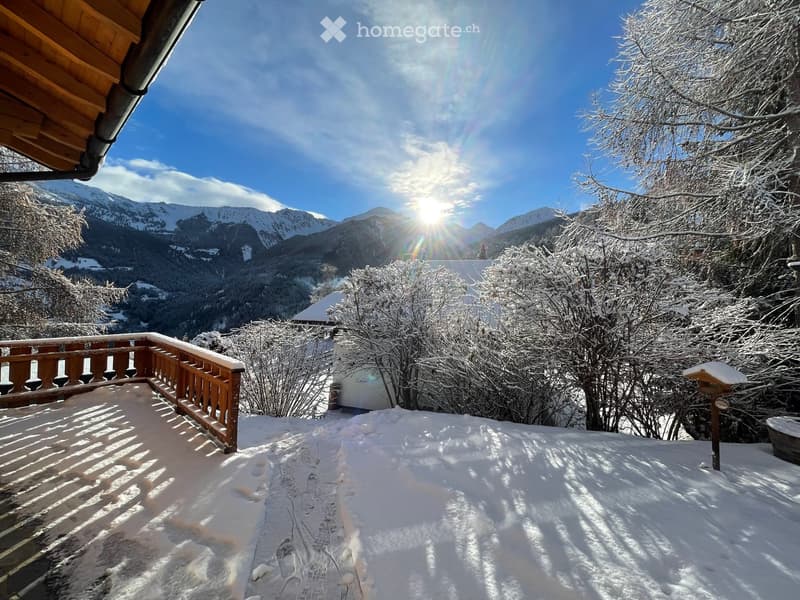 Snow-covered balcony with wooden railing, overlooking a snowy mountain landscape and trees.