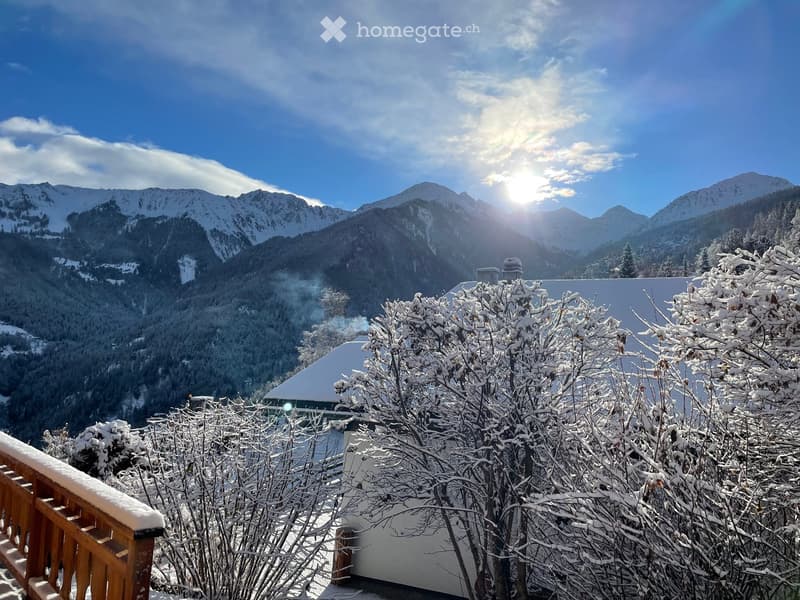 Snow-covered balcony overlooking a mountain range with snow-covered trees and a clear blue sky.
