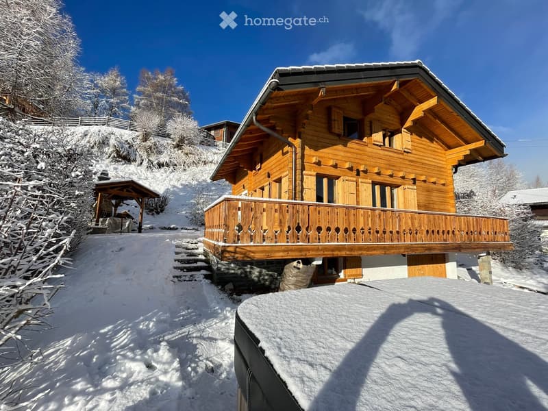 A large wooden chalet with a balcony, situated in a snowy mountain landscape with surrounding trees.