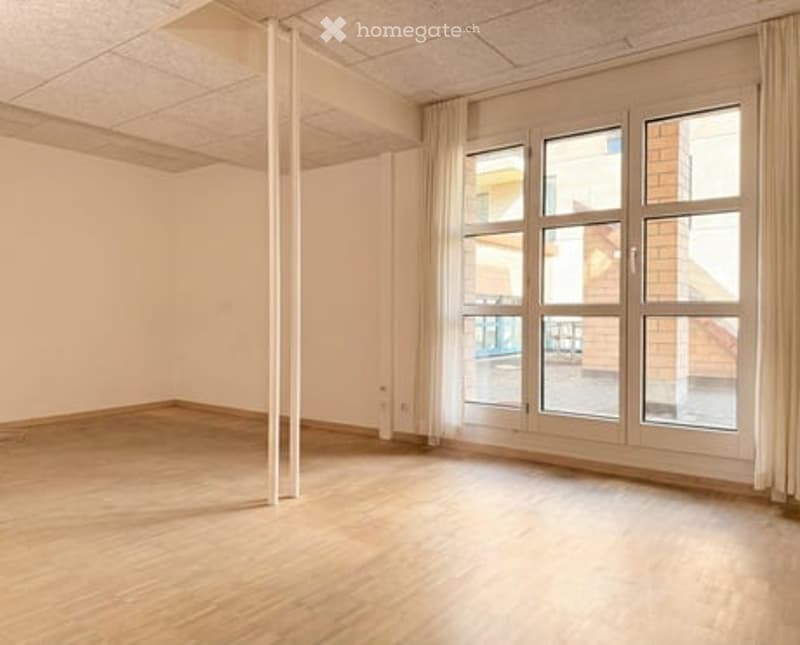 Empty room with wooden flooring, white walls, and large windows with curtains. No furniture or equipment present.