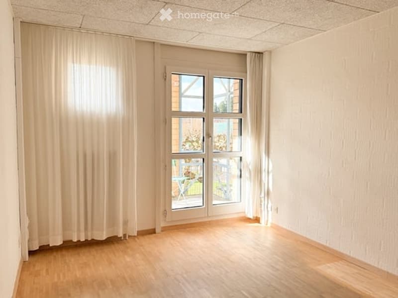Empty room with wooden flooring, white walls, large window with white curtains, and a glass door leading to a balcony.