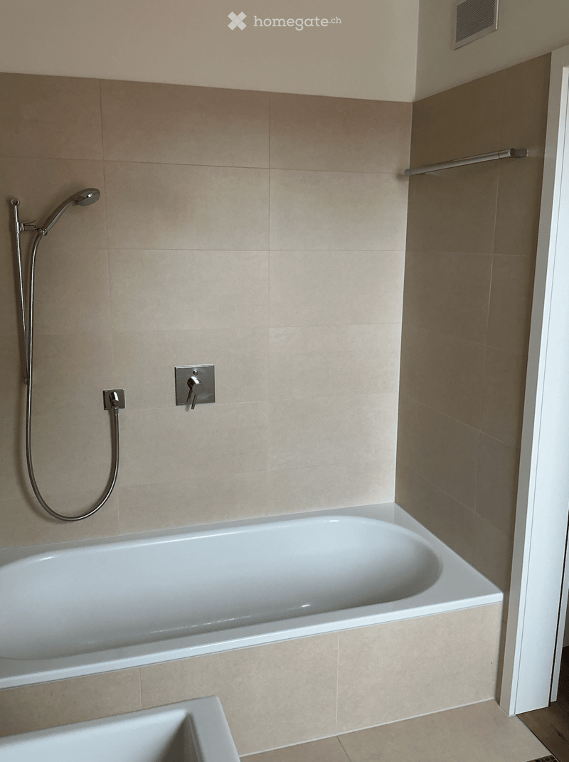 Modern bathroom with a white bathtub, beige tiled walls, and a handheld showerhead mounted on the wall.