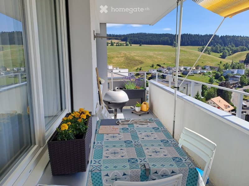 Balcony with a scenic view, featuring a table with a blue and white patterned tablecloth, chairs, a grill, and flower pots.