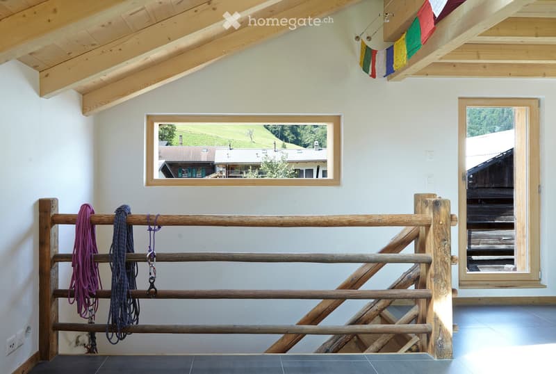 Bright hallway with wooden beams and exposed ceiling, featuring a large window showing a scenic view, a wooden railing with ropes, and colorful flags hanging from the ceiling.