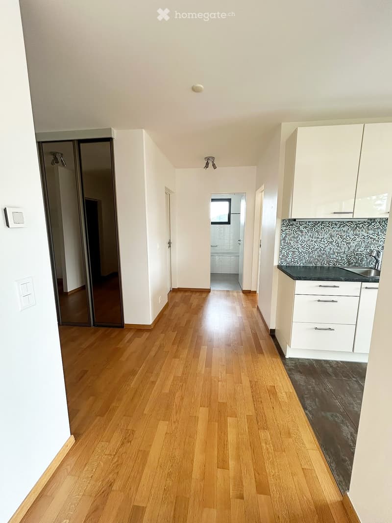 Empty hallway with wooden flooring, white walls, and a small kitchen area visible on the right side.