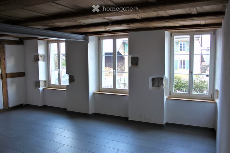 Empty room with exposed wooden beams on the ceiling, tiled floor, and large windows with wooden sills. The walls are white with some exposed stone patches.