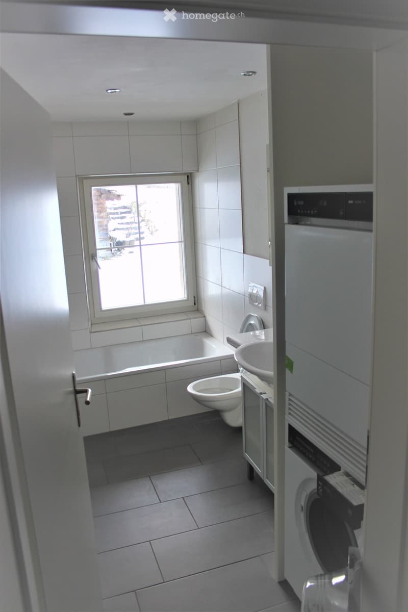 Modern bathroom with white tiles, bathtub, toilet, sink, and built-in washing machine. Features a window with blinds and a door.
