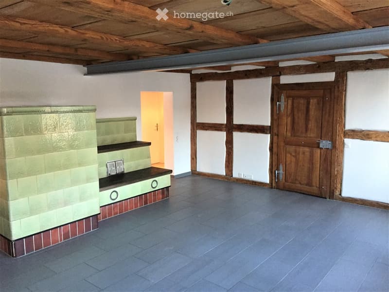Empty room with exposed wooden beams on the ceiling, tiled floor, and a wooden door leading to another space.