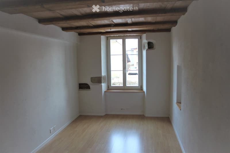 Empty room with wooden flooring, white walls, and a large window with a view of the outside. The ceiling features exposed wooden beams.