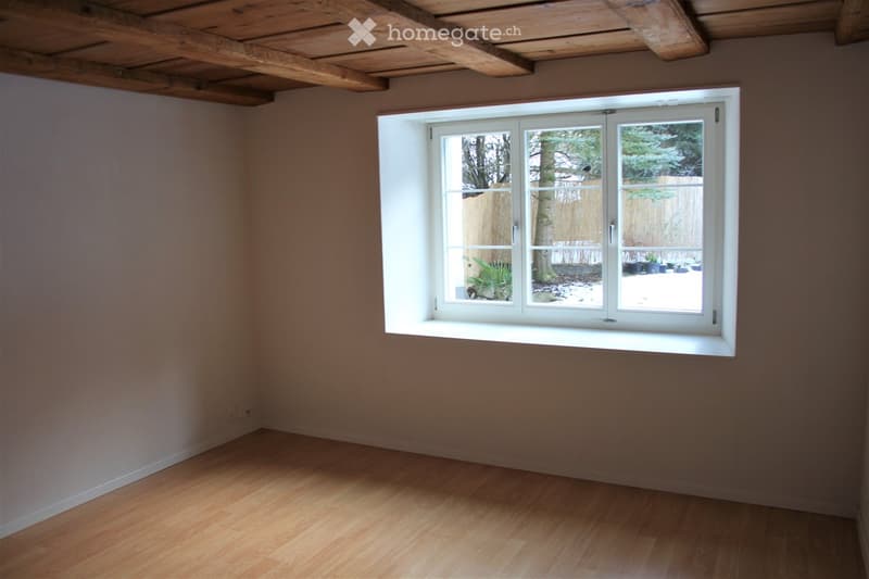 Empty room with wooden flooring, exposed wooden ceiling beams, and a large white-framed window showing a view of a fenced yard with snow and some plants.