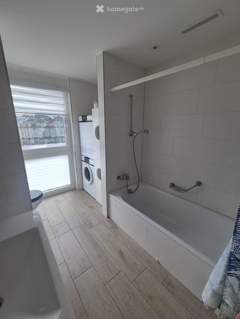 Modern bathroom with a bathtub, showerhead, faucet, washing machine, and toilet. Features wooden flooring and large windows with blinds.