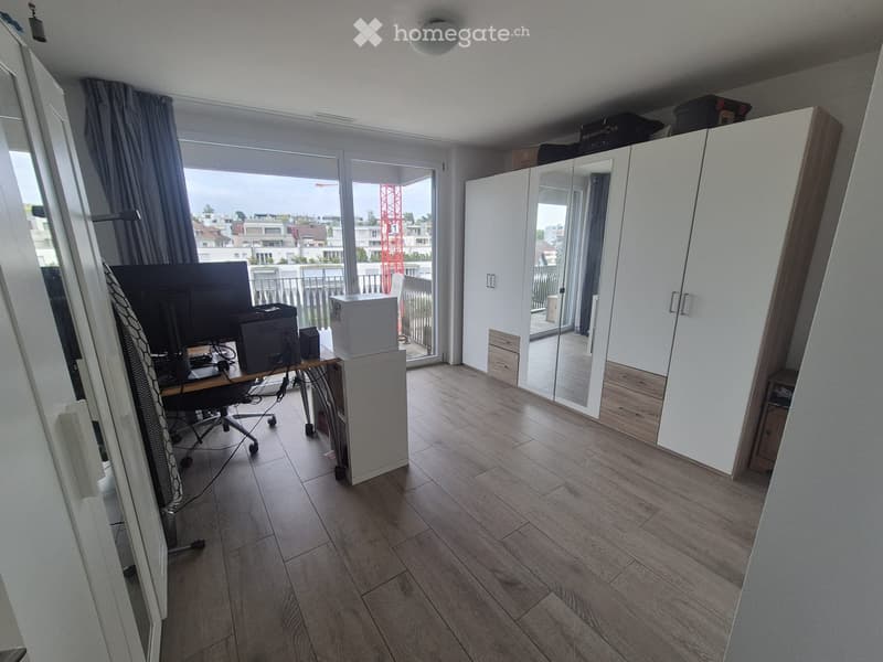 A furnished room with a wooden floor, a desk with a chair, a screen, a printer, and a large white cabinet. There is a balcony with a view of the city.